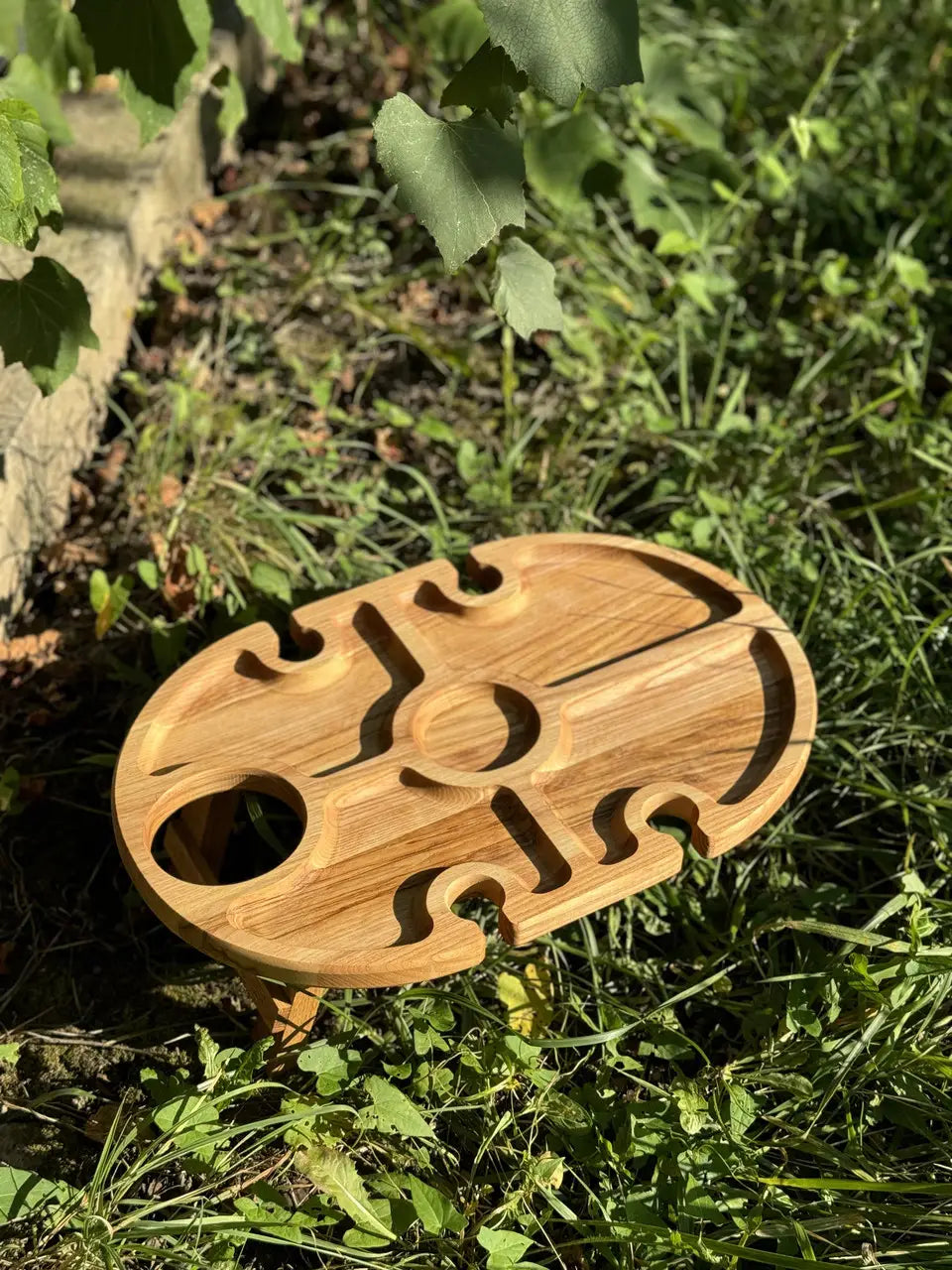Empty oval wooden wine table on grass with carved slots for glasses and bottle.