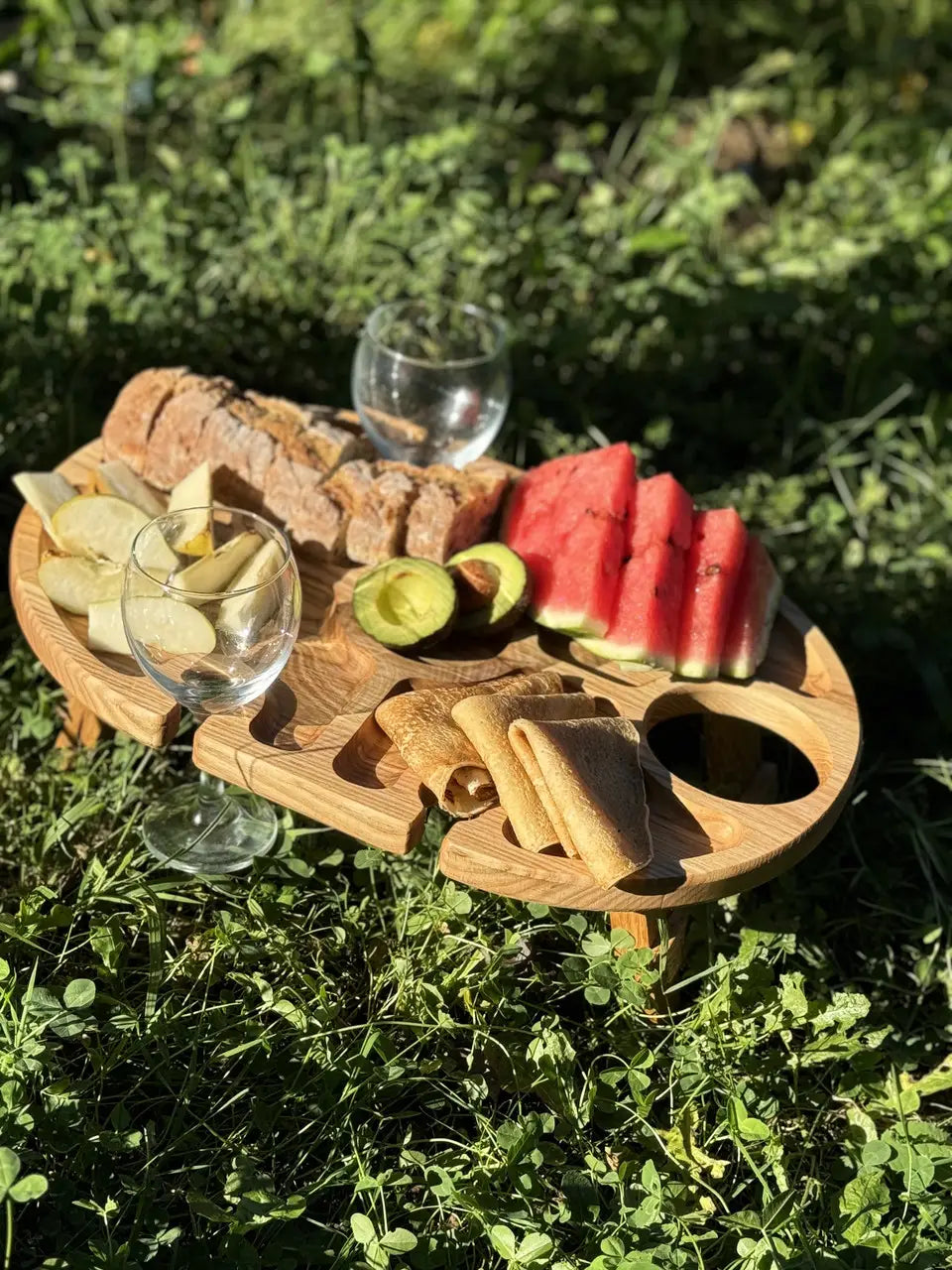 Handmade wooden table with cutouts for wine glasses, served with fresh picnic snacks on the grass.