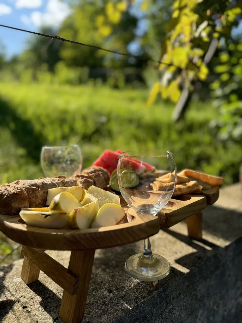 Rustic wine picnic table with fruit, bread, and empty glasses set on outdoor stone surface.