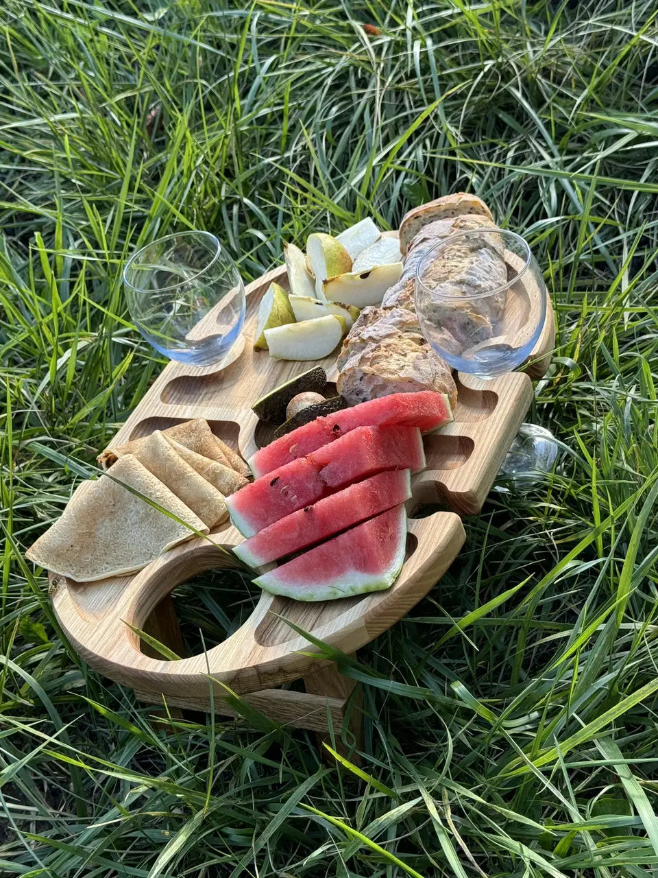 Top view of wooden wine table on grass, filled with watermelon, crepes, pear, bread, and two glasses.