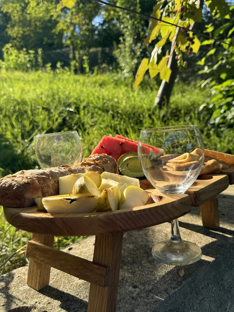 Close-up of wooden picnic table with wine glasses, pear slices, watermelon, avocado, and bread outdoors.