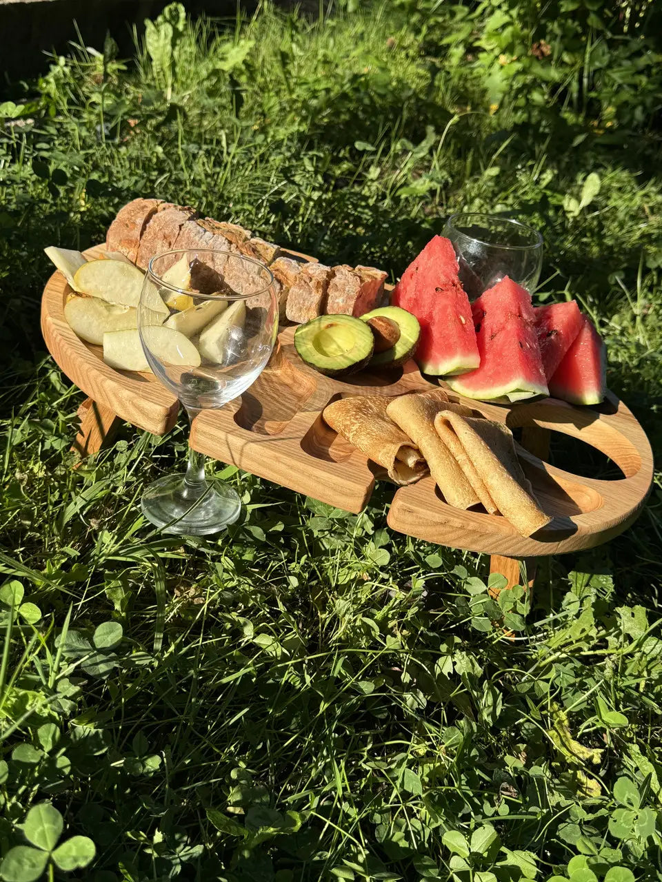 Wooden wine picnic table in nature with food, wine glasses, and greenery in the background.