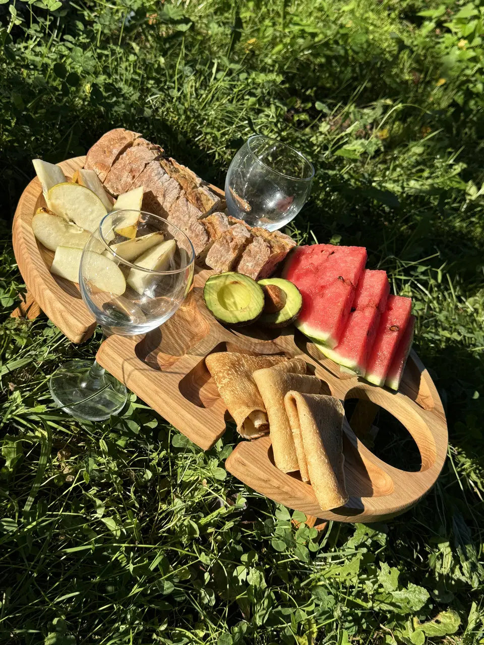 Overhead shot of picnic setup with wooden wine tray, fruits, glasses, avocado, and pancakes.
