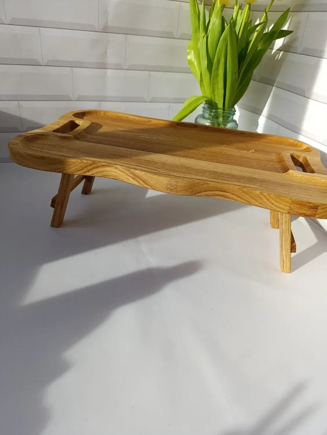 Natural oak breakfast tray table with curved design and cut-out handles, photographed in soft sunlight.