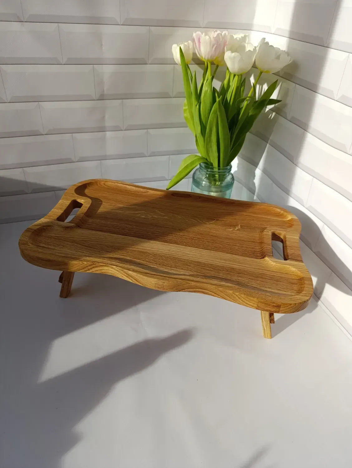 Handcrafted wooden serving tray table with wave-shaped edges, placed beside a vase of tulips.