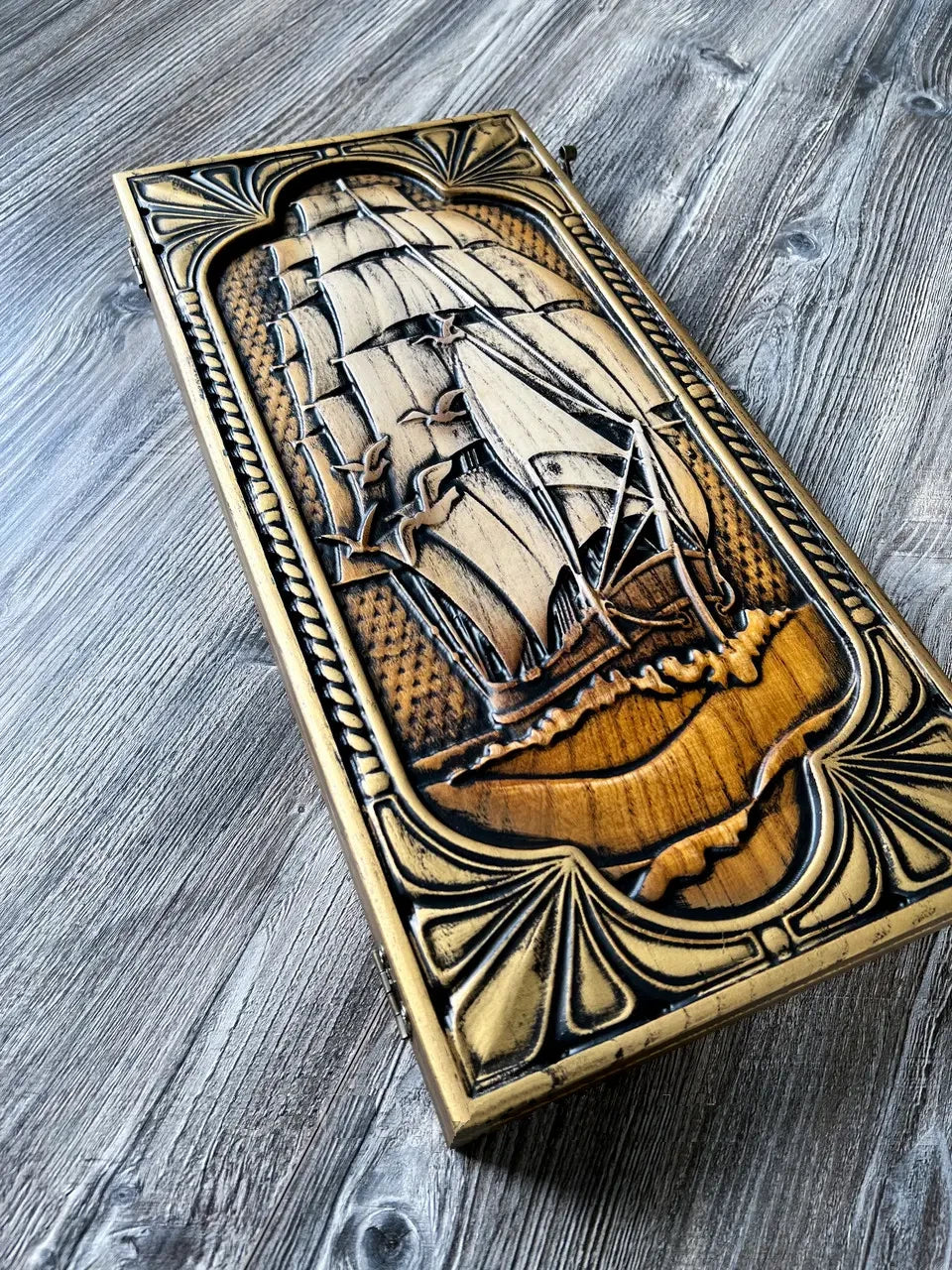 Handcrafted wooden backgammon case with carved sailing ship on textured wood background.