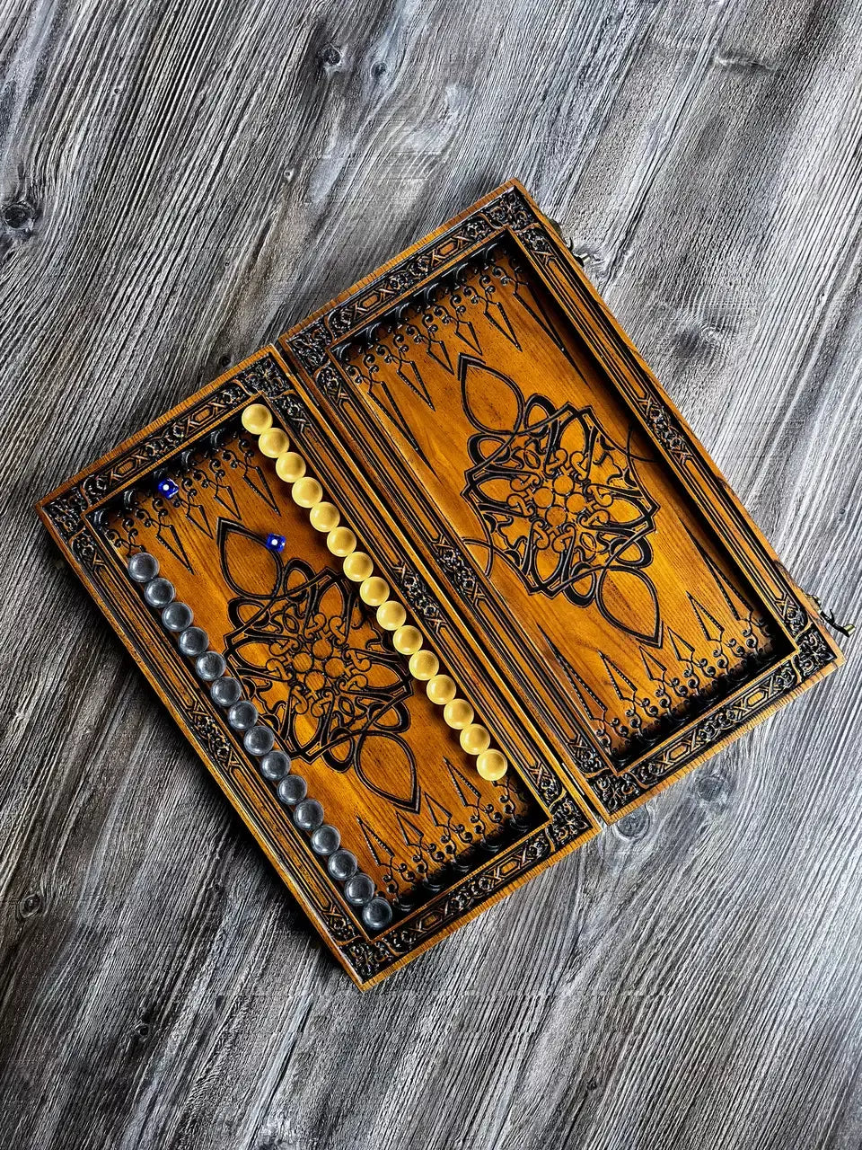 Open wooden backgammon board with oriental floral engraving, game chips, and dice on a rustic wooden surface.